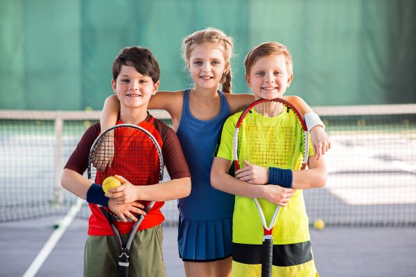 We like tennis. Happy girl is embracing two boys while standing on playground. They are holding tennis racquets and smiling. Portrait